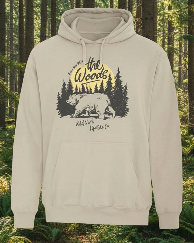 A beige unisex hoodie with a graphic print of a bear and trees on the front and the text 'The Woods' above the image.