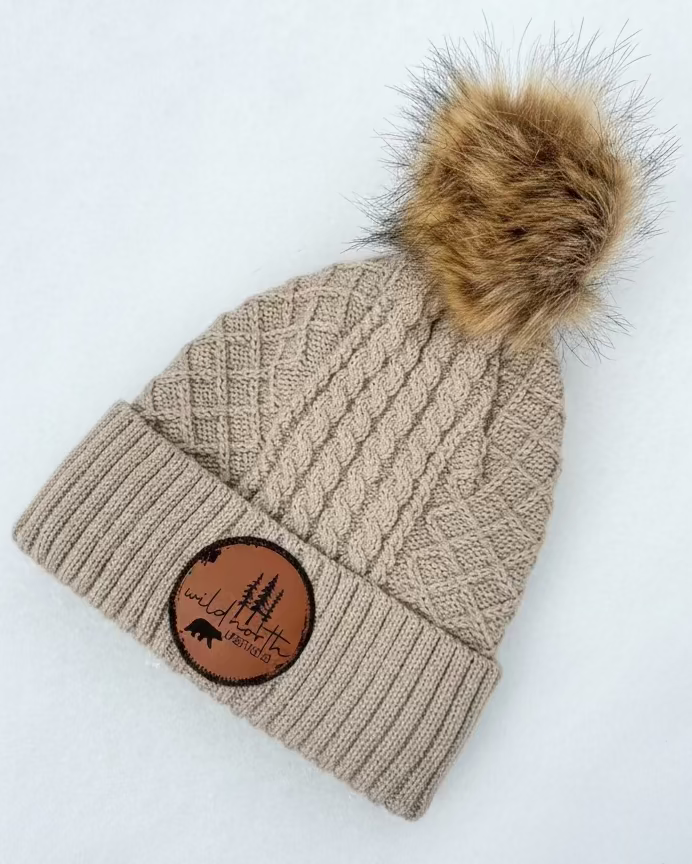Beige knit beanie with fur pom-pom and leather patch on a white background