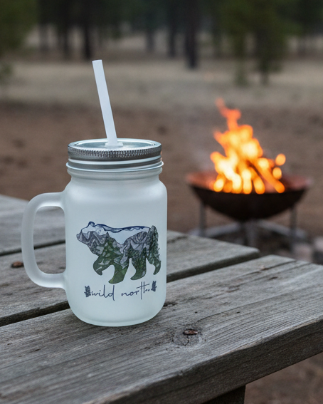 Mason jar with a straw on a wooden table near a campfire and green chair in a forest setting