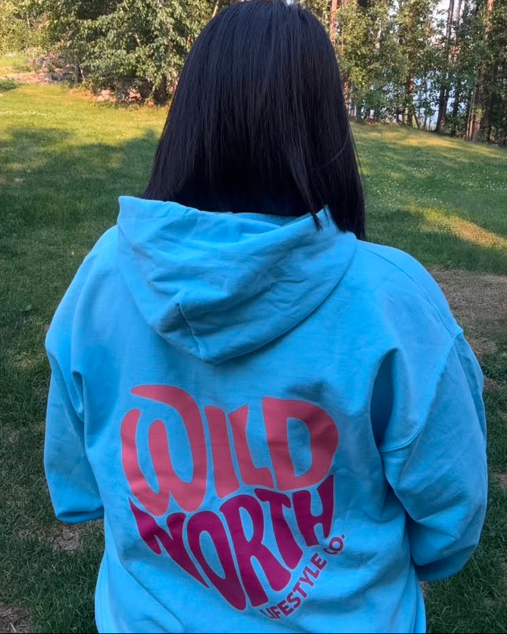 A person viewed from behind wearing a light blue hoodie with a large pink 'WILD NORTH LIFESTYLE CO' graphic on the back, standing outdoors on a grassy lawn with trees in the background.