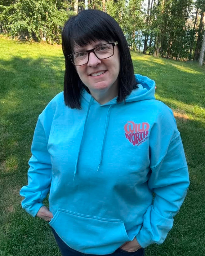 A woman with dark hair and glasses wearing a light blue hoodie with a pink 'Wild North' graphic on the left chest, standing outdoors on a grassy lawn with trees in the background.