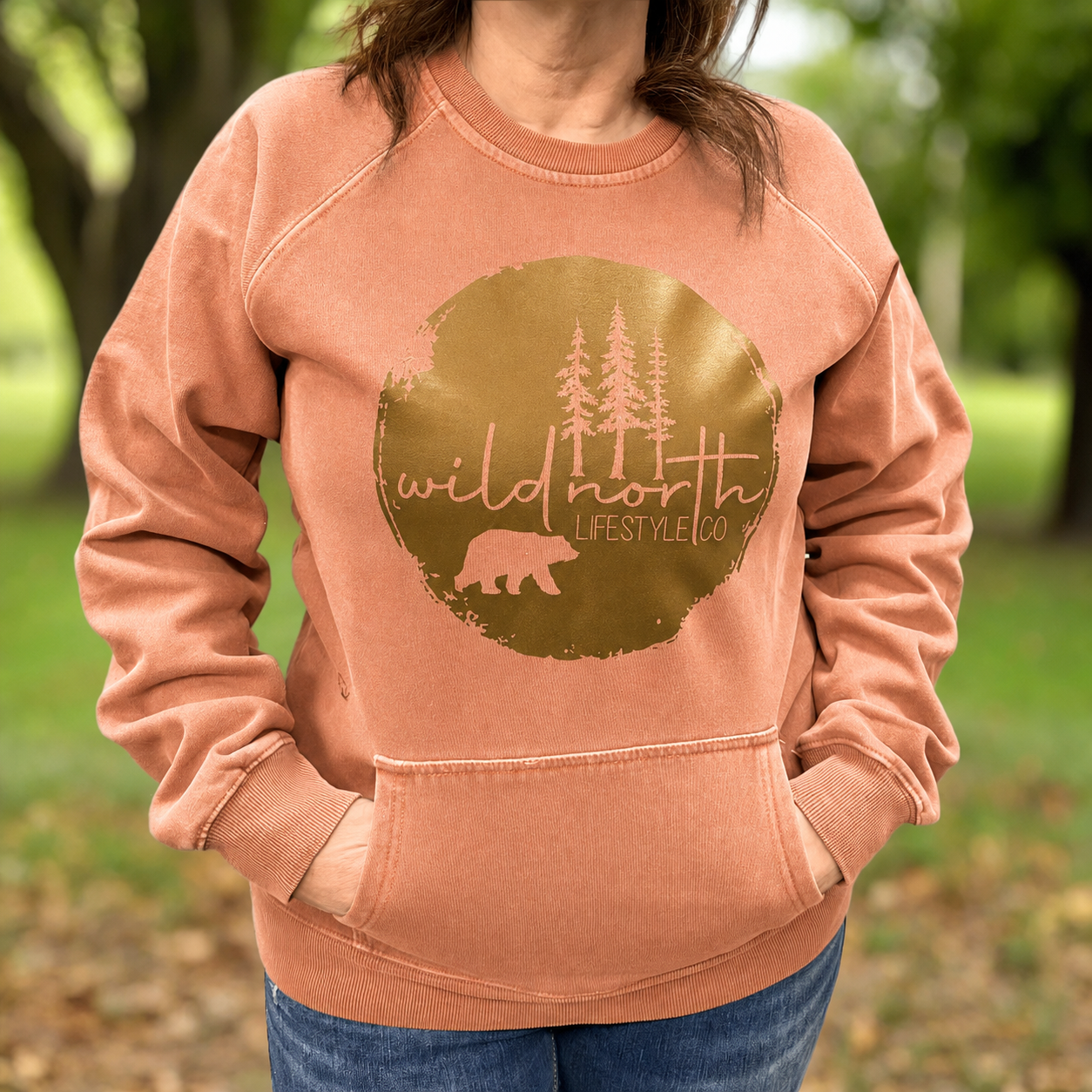 A person wearing a peach crewneck sweatshirt with a large gold circular graphic on the chest that reads 'wild north LIFESTYLE CO' with pine trees and a bear silhouette, photographed outdoors in a blurred park setting.